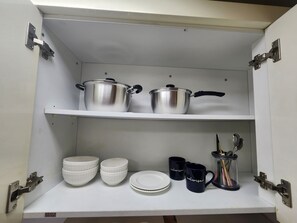 Ein Küchenschrank mit Edelstahl-Kochtöpfen, weißen Schüsseln, Tellern und einer Kaffeemaschine.