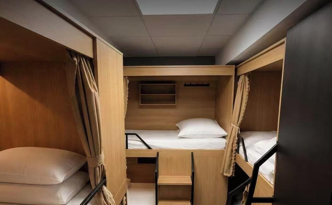 Quarto pequeno com beliches, uma prateleira e cortinas.