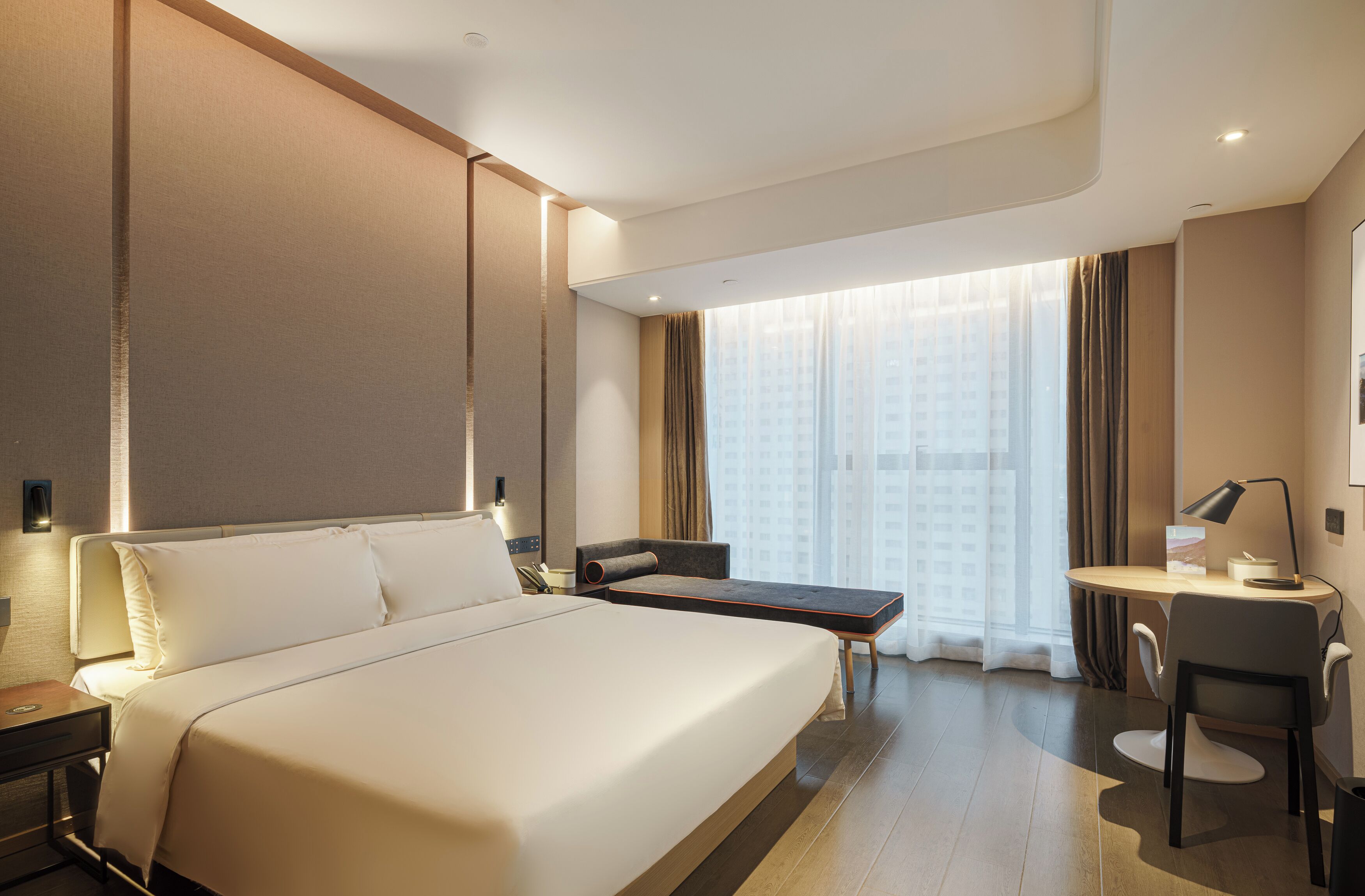 A hotel room with a large bed, a desk, a chair, and a view of a building through the window.