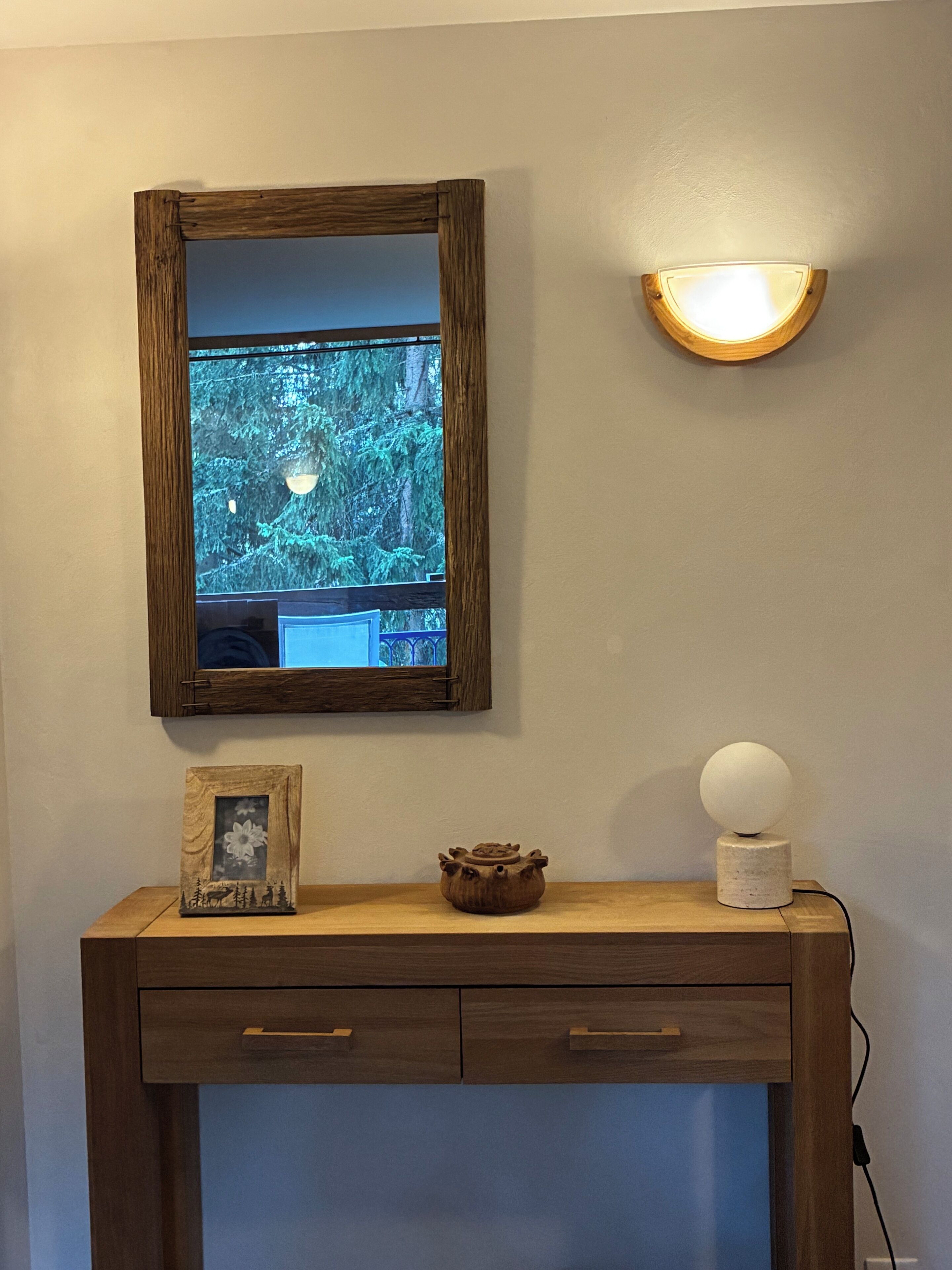Una consola de madera con un cuadro enmarco, una maceta de cerámica y un aplique de pared.