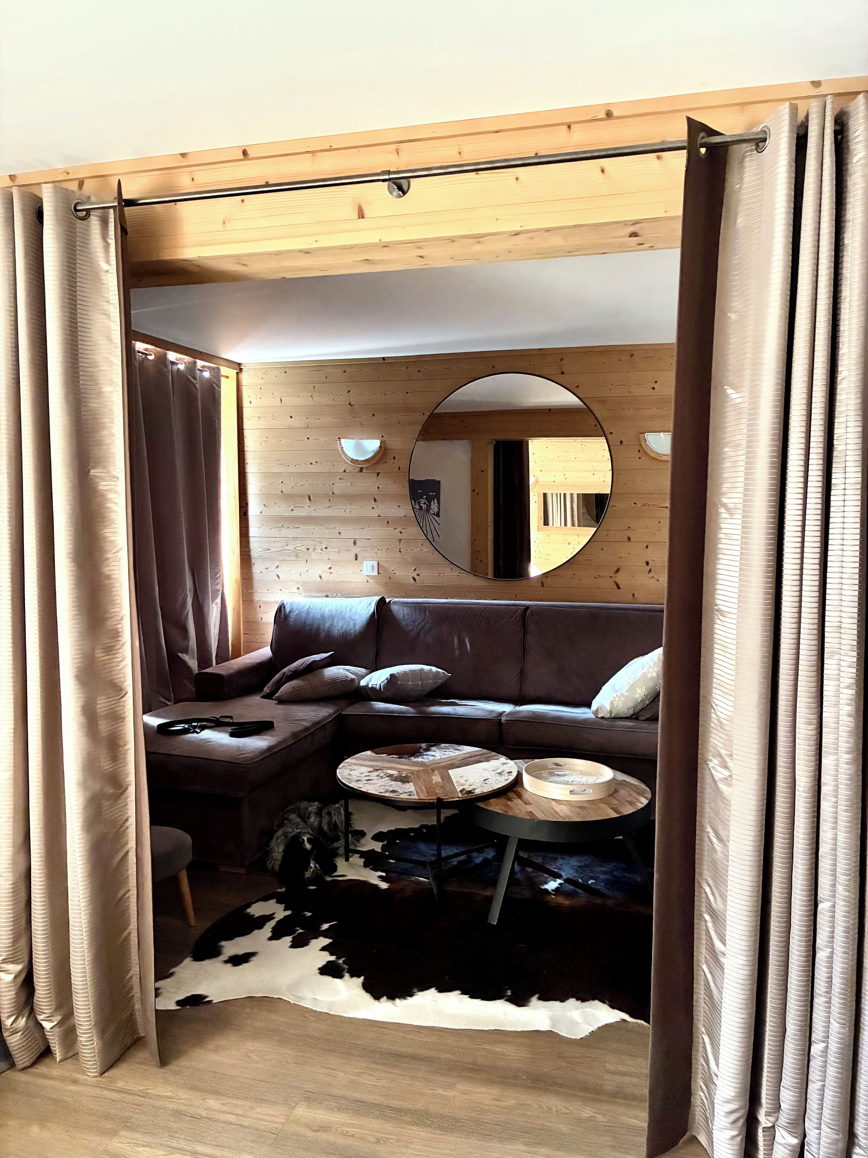 A cozy living room with a brown leather sofa, a round coffee table, and a cowhide rug. A wooden ceiling and a wall-mounted mirror are also visible.