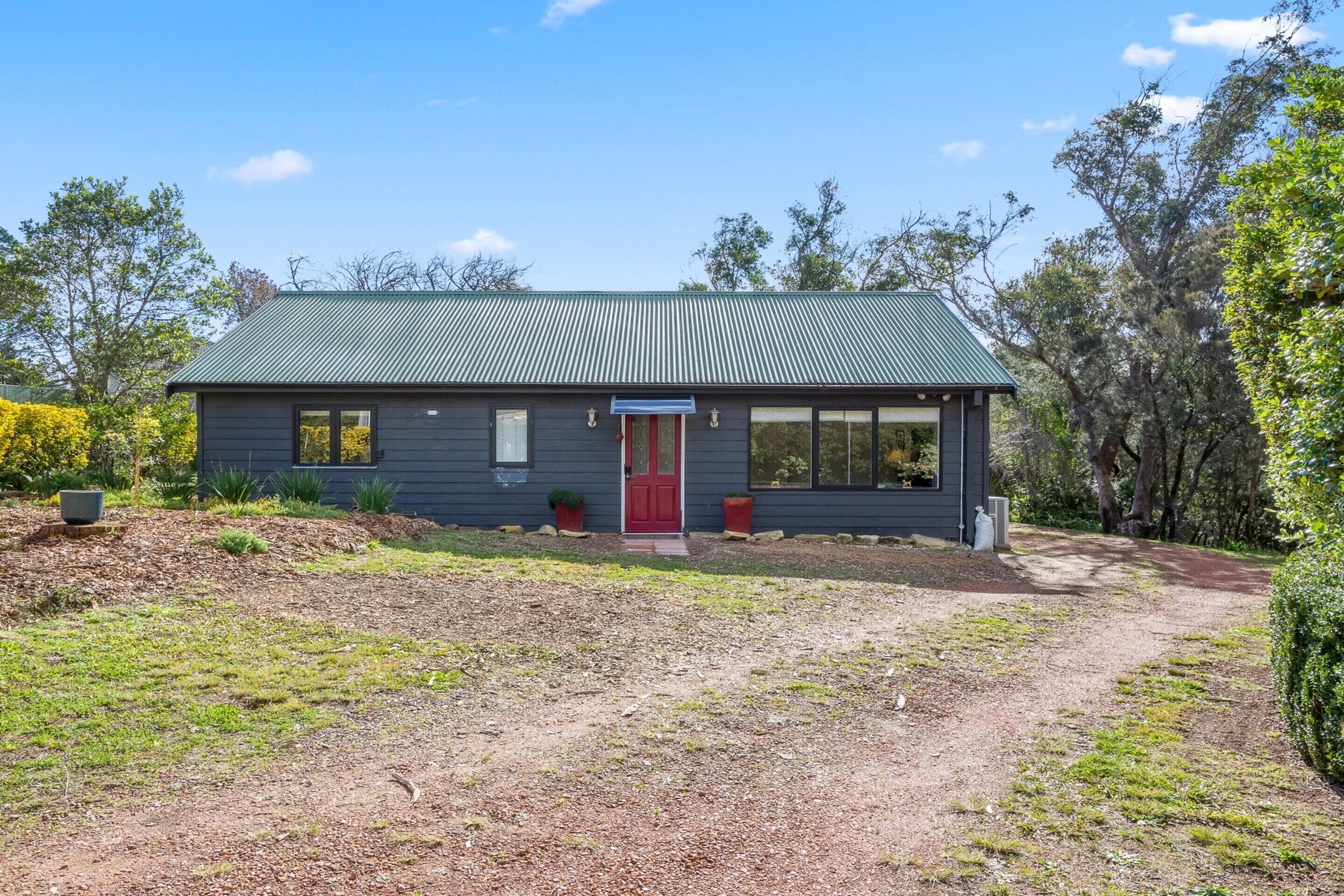 A single-story house with a dark-colored exterior, a red door, and a green metal roof.