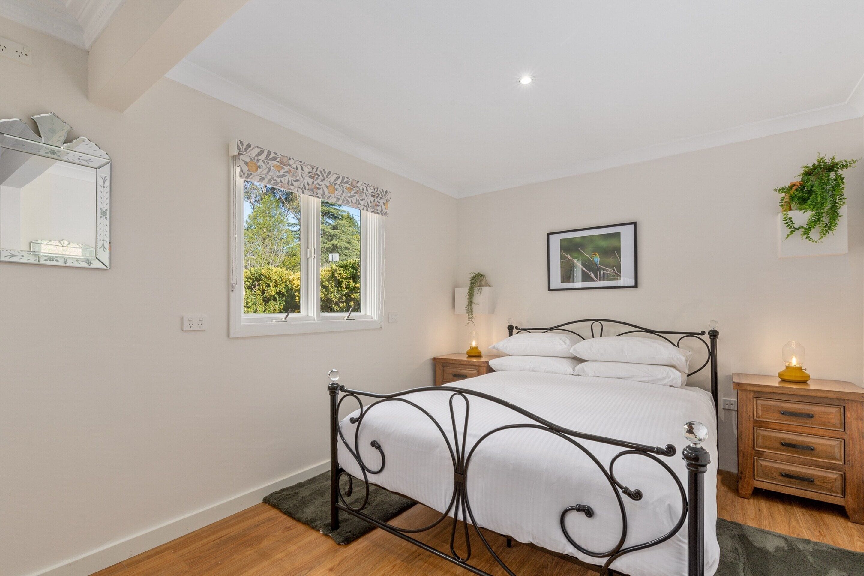 A bedroom with a bed, bedside tables, a mirror, a window with curtains, a wall-mounted picture, and a plant.