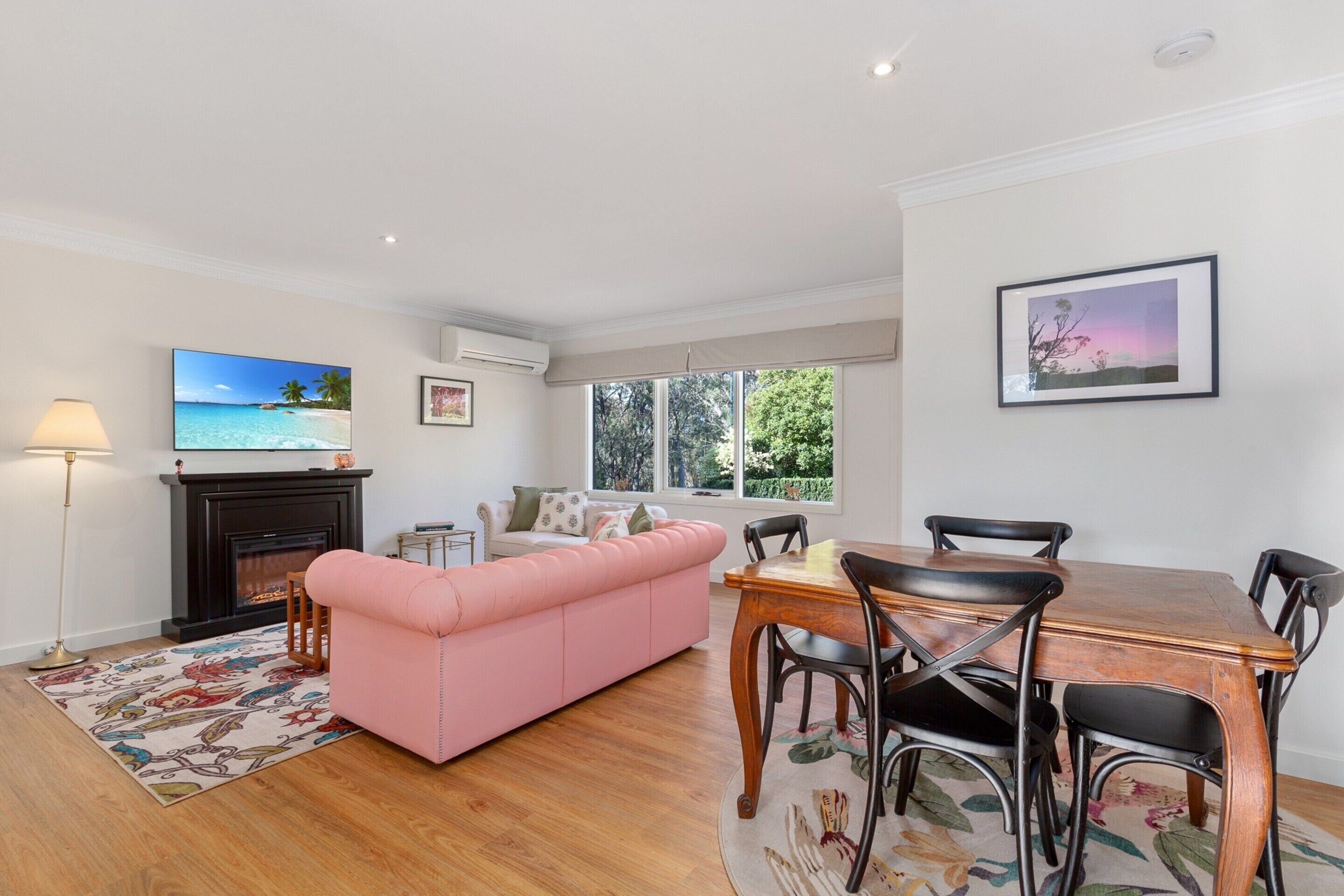 A living room with a pink sofa, a fireplace, a television, and a dining area with a table and chairs.