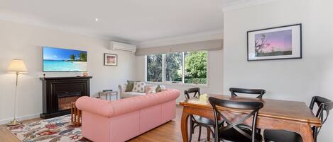A living room with a pink sofa, a fireplace, a television, and a dining area with a table and chairs.
