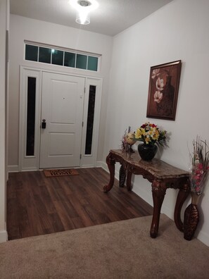 Ein Flur mit Holzboden, einer Fußmatte, einer Tür mit Frostglas-Elementen, einem gerahmten Bild an der Wand und einem Tisch mit Blumen in einer Vase.