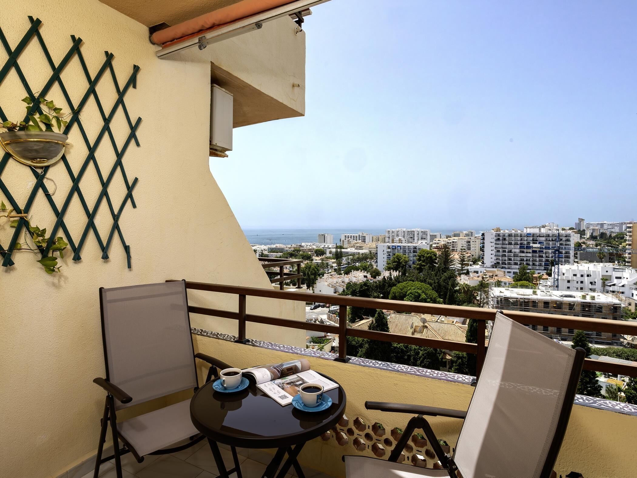 A balcony with a table and chairs, a view of the cityscape, and a lattice wall with plants.