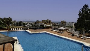 Piscina con sillones de descanso y sombrillas, rodeada de edificios bajo un cielo despejado.