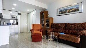 Una sala de estar moderna con un sofá, un sillón y una mesa de centro, una zona de cocina con microondas y una mesa de comedor.