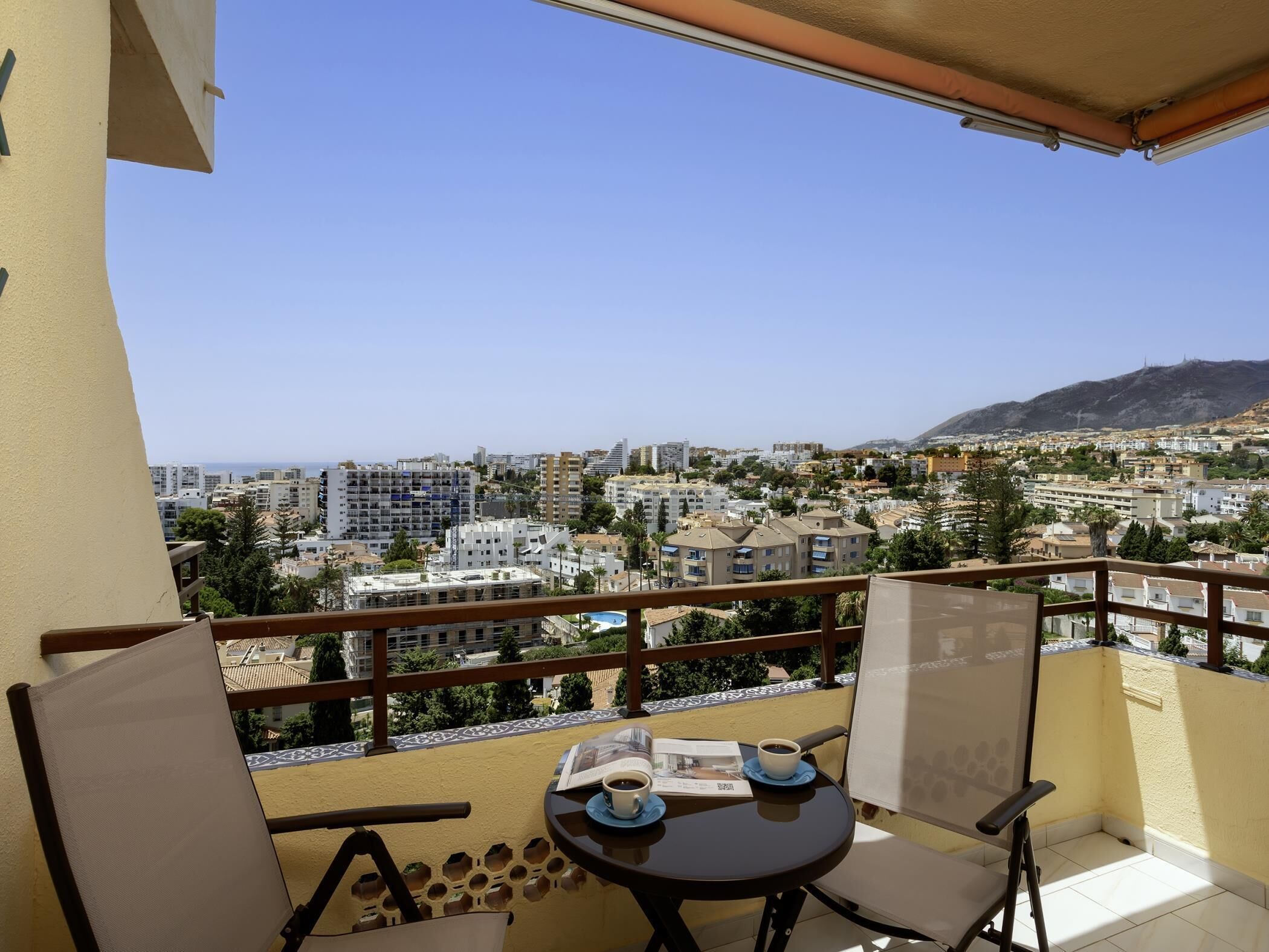 A balcony with a table, chairs, and a view of a cityscape with buildings and a mountain.