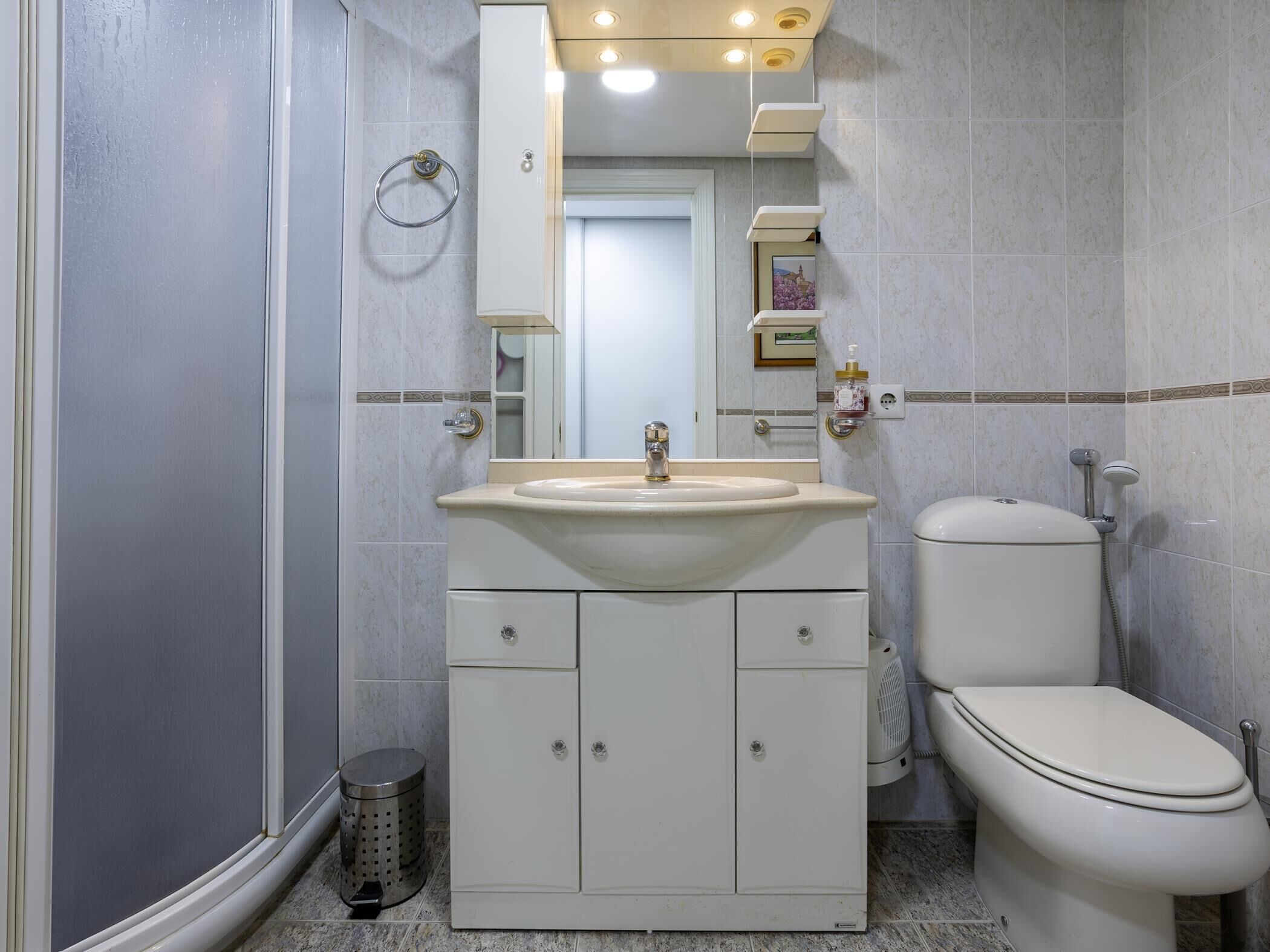 Baño con lavamanos, espejo, inodoro y un cubo de basura.