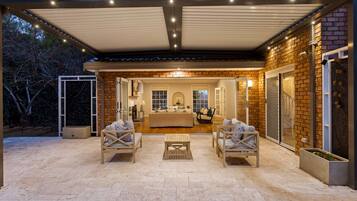 An outdoor patio area with a covered seating arrangement, a kitchen visible through an open door, and a brick exterior wall.