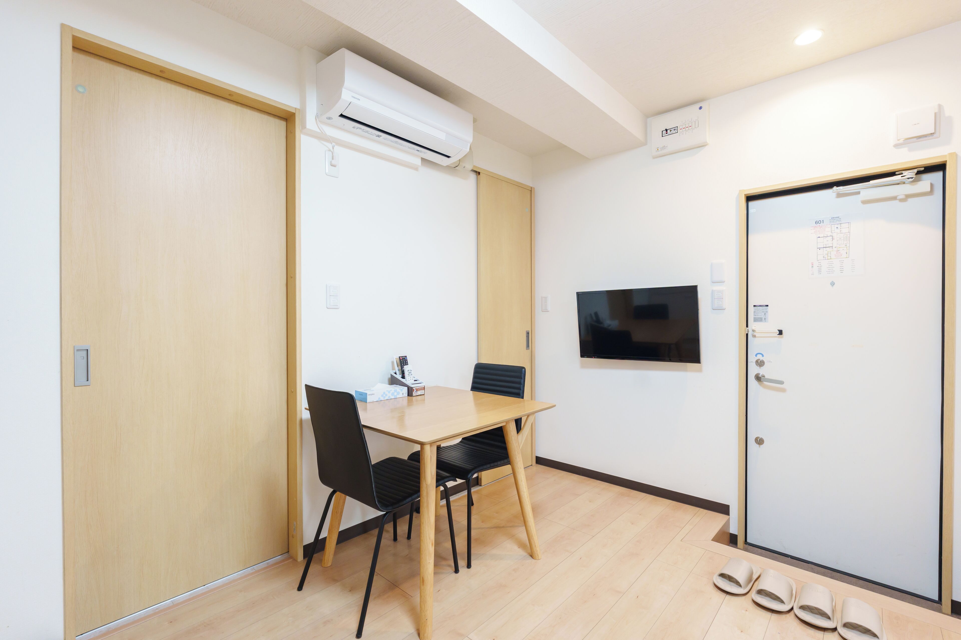 A hotel room with a wooden table, two chairs, a television mounted on the wall, and a door with a room number sign.