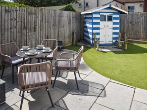 Eine Terrasse mit Korbstühlen und einem Esstisch, ein kleines Gartenhaus mit blauen und weißen Streifen sowie ein Holzzaun.