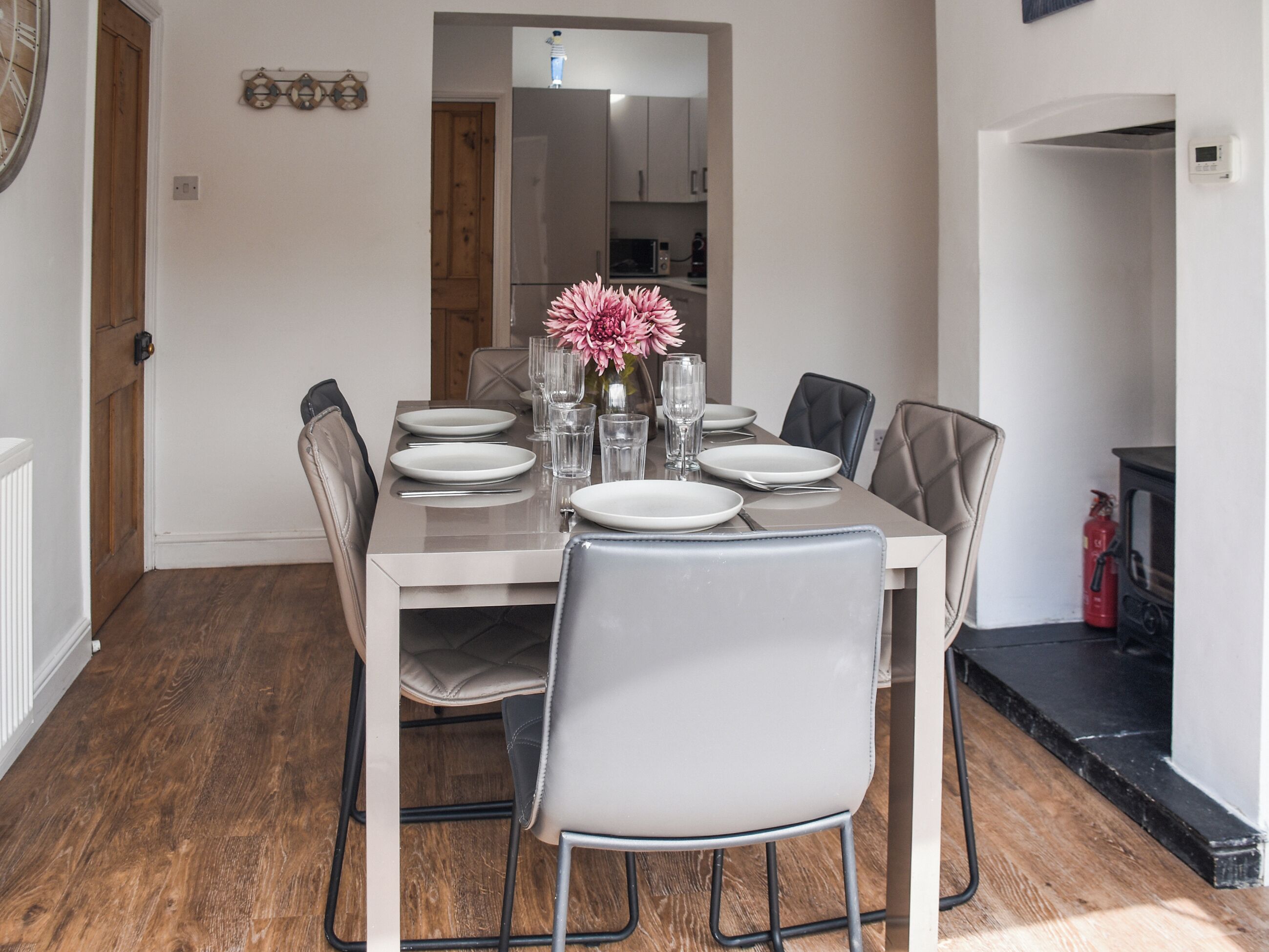 A dining area with a table set for six, chairs, a vase with flowers, and a kitchen in the background.