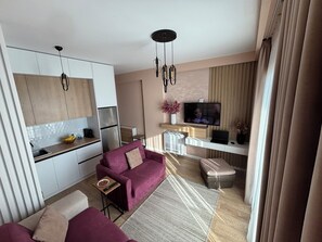 Ein modernes Wohnzimmer mit einer Couch, einem Couchtisch und einem Fernseher. Es gibt eine Küchenzeile mit Schränken und einem Kühlschrank.