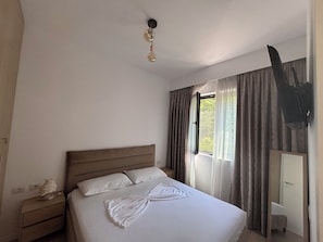 Ein Hotelzimmer mit einem Einzelbett, einem Nachttisch, einem Fenster mit Vorhängen und einem an der Wand befestigten Fernseher.