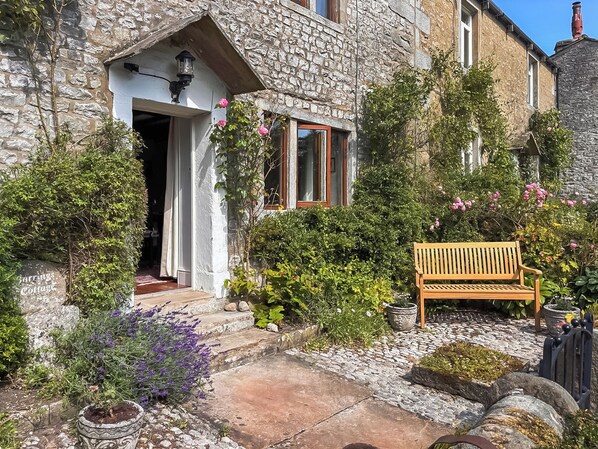 A stone cottage with a wooden bench, a stone pathway, and a variety of plants and flowers.