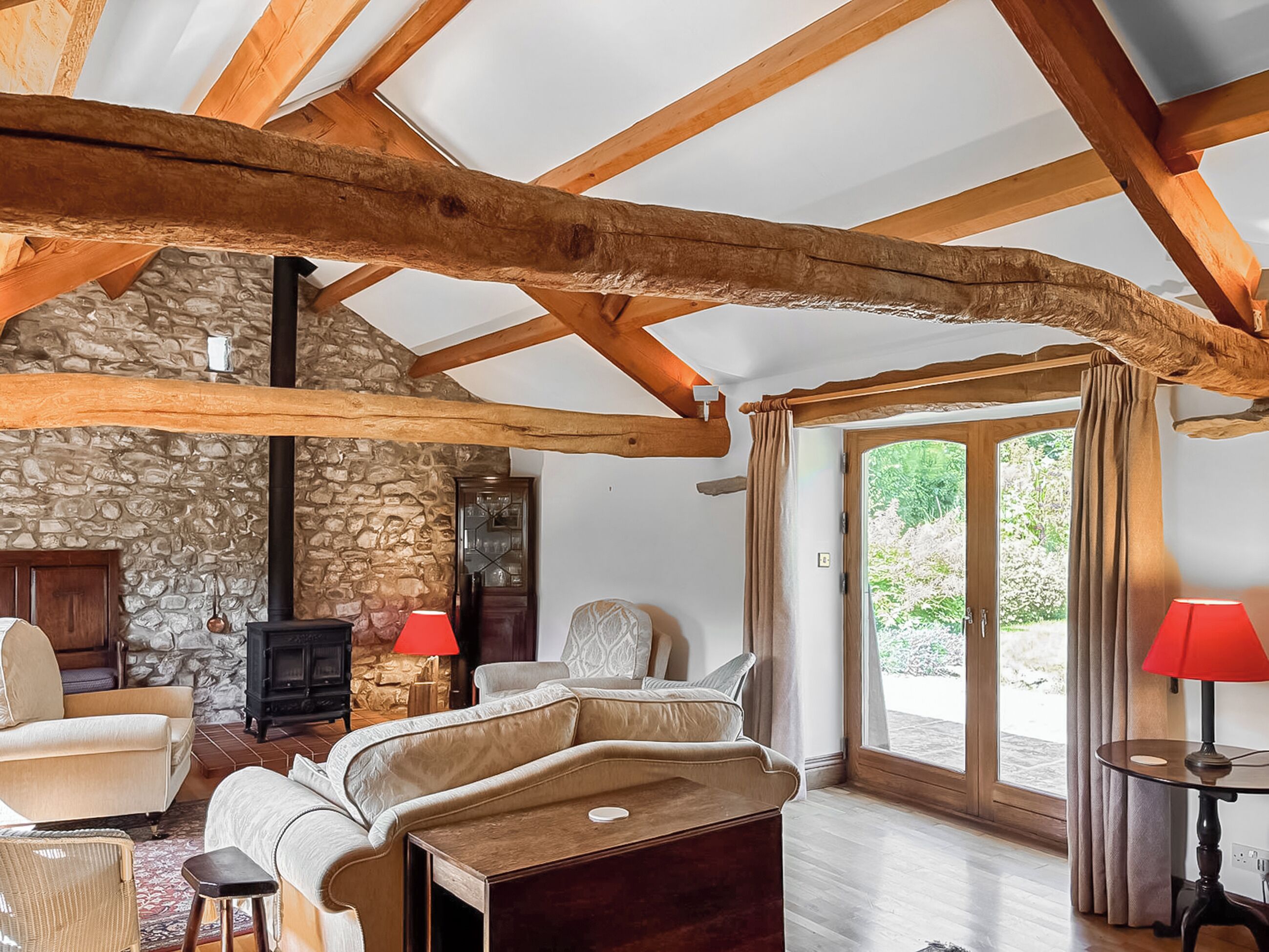 A cozy room with a stone wall, wooden beams, a fireplace, a sofa, and a table with a lamp.