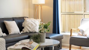 Ett vardagsrum med en soffa, ett soffbord med ett tidningsnummer, en golvlampa och ett fönster med gardiner.