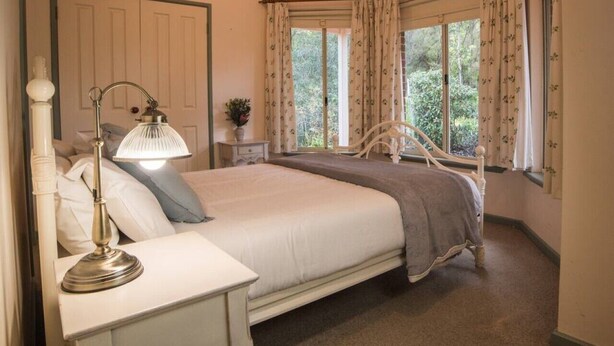 A bedroom with a bed, bedside table, lamp, and curtains. - Whispering Karri Retreat | 5-Acre Hideaway (Shadforth)