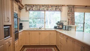 A kitchen with wooden cabinets, a window with curtains, and a view of the outdoors. - Whispering Karri Retreat | 5-Acre Hideaway (Shadforth)