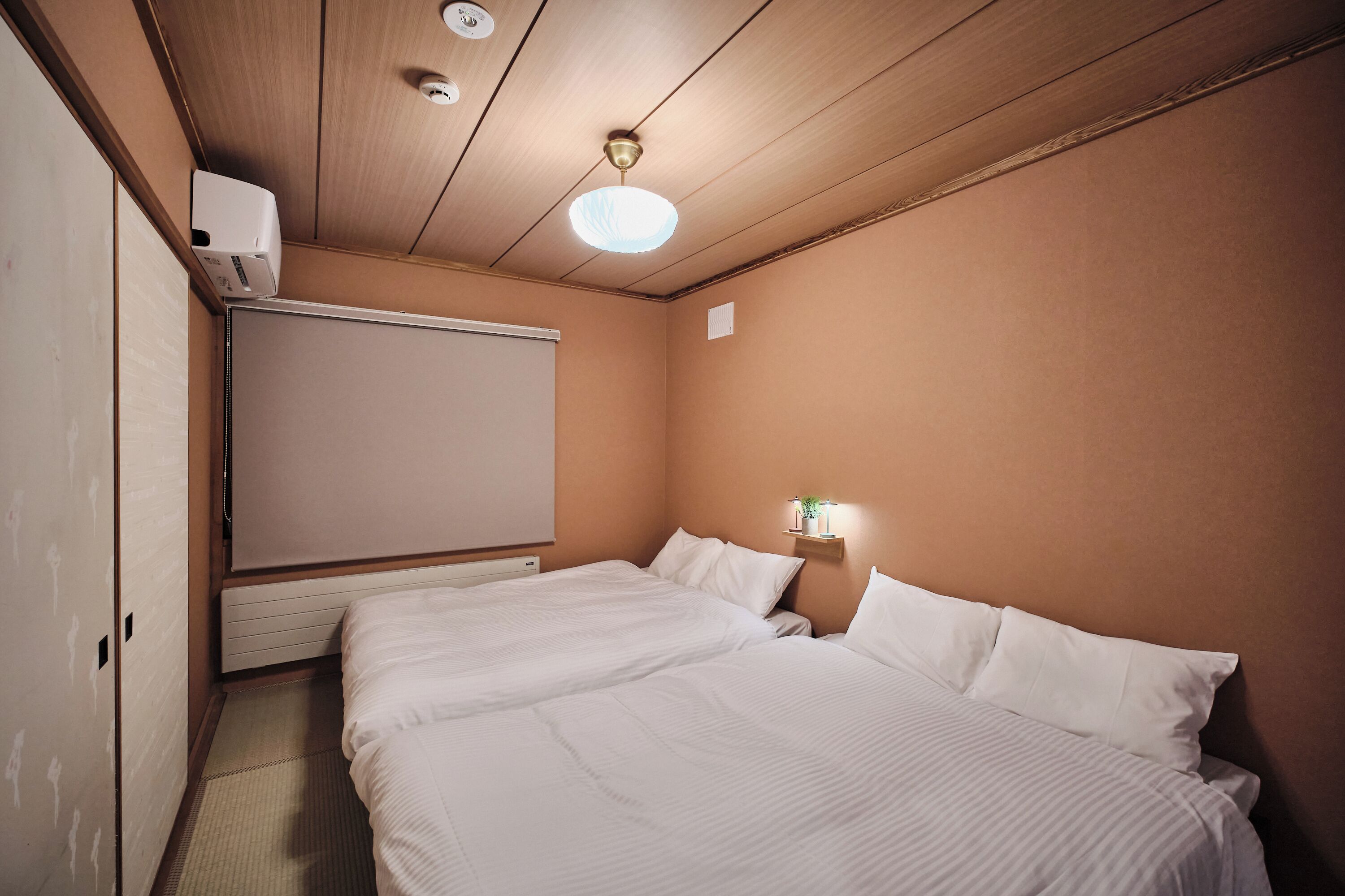 A hotel room with a bed, a ceiling fan, a wall-mounted heater, and a small bedside table with a lamp.