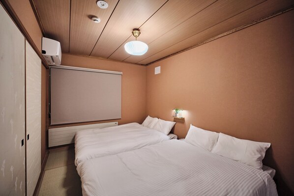 A hotel room with a bed, a ceiling fan, a wall-mounted heater, and a small bedside table with a lamp.
