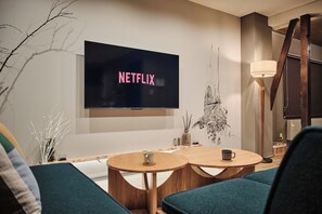 A modern living room with a flat-screen TV displaying 'NETFLIX' on the wall, two round wooden tables, and a floor lamp.