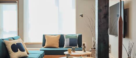 A modern living room with a teal sofa, a round wooden coffee table, and a large window providing natural light.