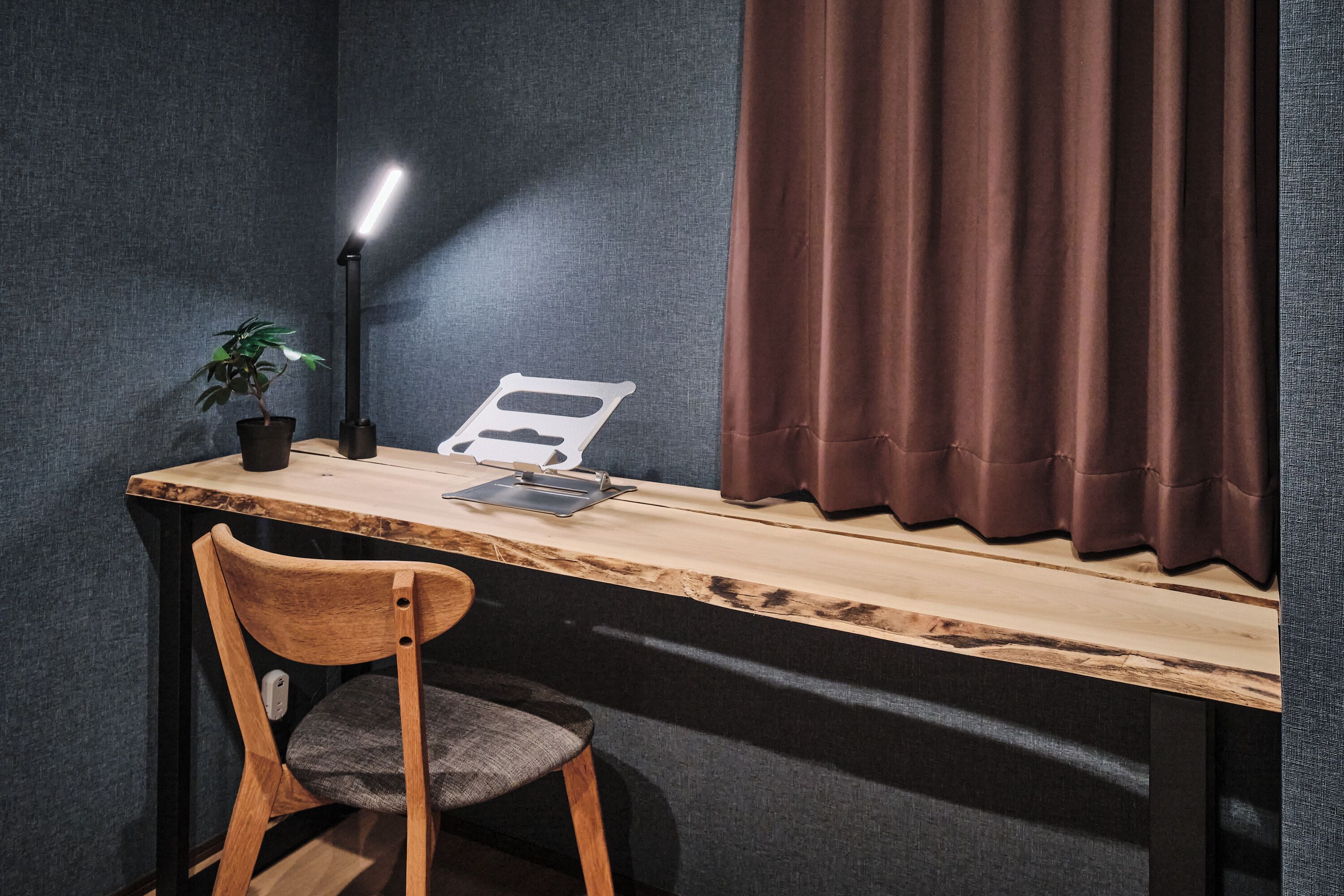 Un escritorio con una lámpara, una planta y una silla en una habitación de paredes oscuras y una ventana con cortinas.