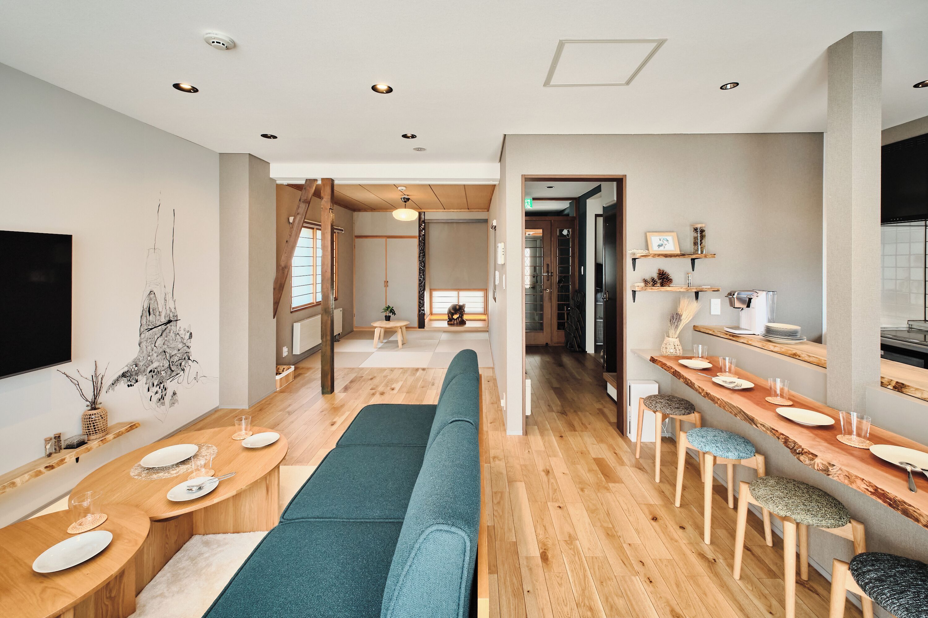Un intérieur moderne avec un long banc en bois, un coin repas avec des tables et des chaises, et un espace cuisine doté d’un comptoir et de tabourets.