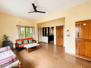A hotel room with a wooden floor, a sofa with cushions, a bed with a floral bedspread, a ceiling fan, and a wooden wardrobe.