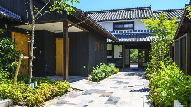 A traditional Japanese building with a tiled roof and wooden walls, a covered walkway, and a garden area with plants and a tree.