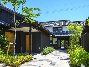 A traditional Japanese building with a tiled roof and wooden walls, a covered walkway, and a garden area with plants and a tree.