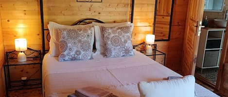 Un dormitorio de cabaña de madera con una cama, dos mesitas de noche con lámparas, una silla de estructura metálica y un armario de madera.
