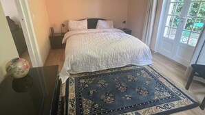 Een slaapkamer met een bed, nachtkastje, lamp, tafel met vaas en uitzicht op groen via een raam.