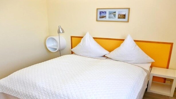 Un letto con una coperta bianca, due cuscini bianchi, un comodino con una lampada e un quadro appeso al muro.