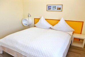 En säng med ett vitt täcke, två vita kuddar, ett sängbord med en lampa och en tavla som hänger på väggen.