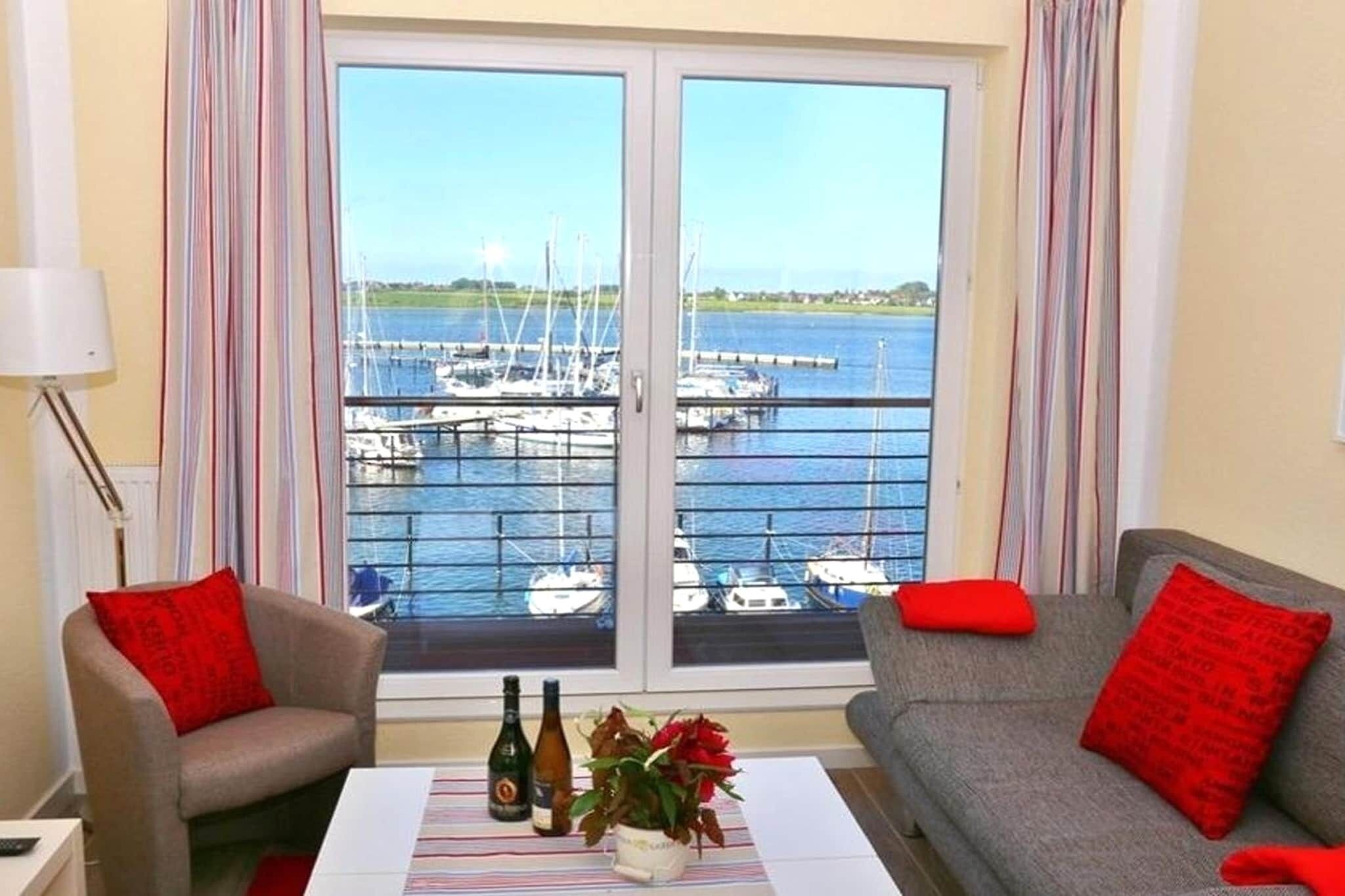 Una habitación con vista a un puerto deportivo, dos sofás con cojines rojos, una mesa con un jarrón de flores y botellas, y una lámpara a la izquierda.