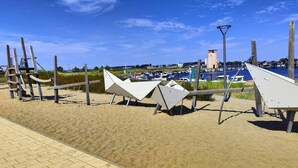 Una spiaggia sabbiosa con una fila di strutture bianche a forma triangolare, un cielo azzurro limpido e un porto turistico con barche sullo sfondo.