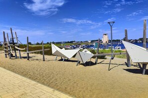 A sandy beach with a row of white triangular structures, a clear blue sky, and a marina with boats in the background. - Apartment Fehmarn-Burgtiefe (Fehmarn OT Burgtiefe-Südstrand)