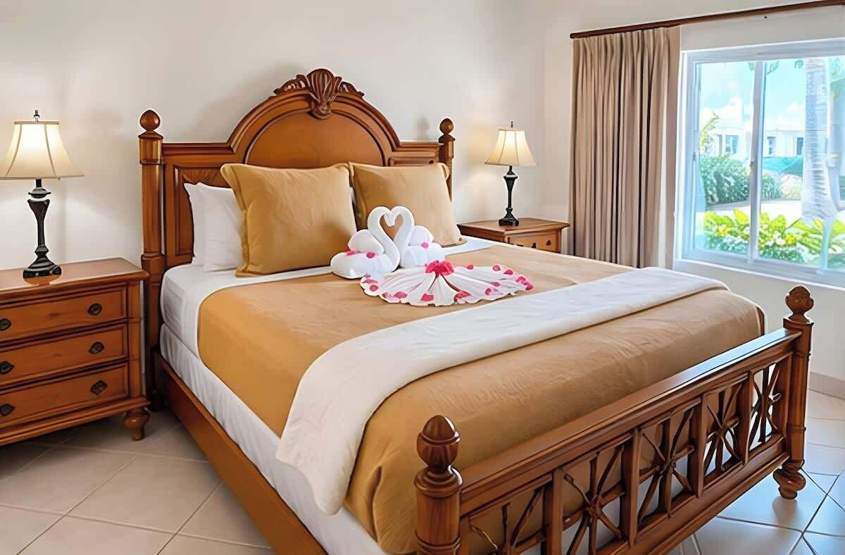 A hotel room with a wooden bed, two bedside tables with lamps, a window with curtains, and a view of greenery outside.