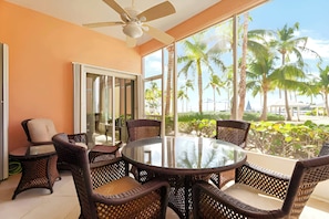A room with a round glass table surrounded by wicker chairs, a ceiling fan, and large windows offering a view of palm trees and a beach. - Kaibo Yacht Club  2 Bdr Beachfront Condo Hot Tub and Pool (Rum Point)