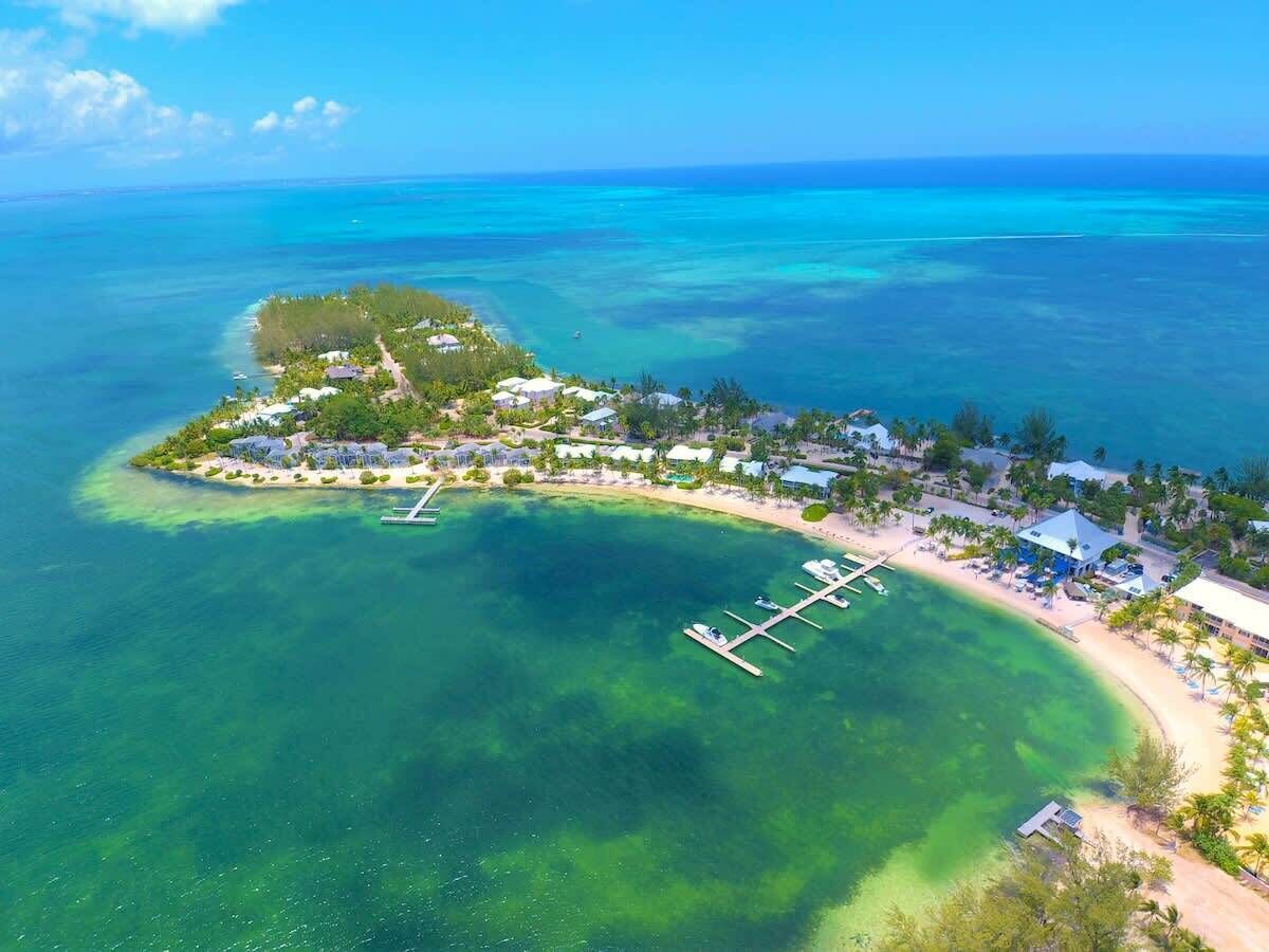 Vue aérienne d’une station balnéaire avec des eaux turquoise cristallines, un port de plaisance et une petite île parsemée de bâtiments et de verdure.