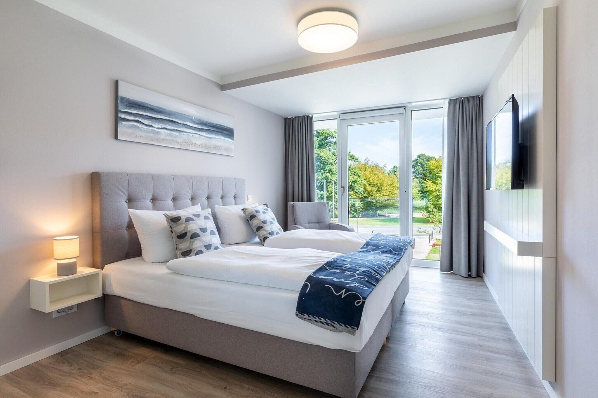 A modern bedroom with a large bed, bedside tables, a flat-screen TV, and a view of the outdoors.