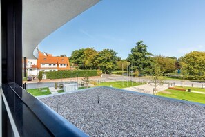 A view from a balcony with a gravel floor, a road with a pedestrian crossing, and a residential area with houses and trees. - Deluxe Baltic Stay für 6 Personen (Travemünde)