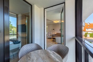 Ein modernes Interieur mit Glastür, die auf einen Balkon führt, einer Sitzgruppe mit gepolsterten Stühlen sowie ein sichtbares Wohnzimmer mit Sofa und Lampe.