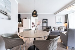A modern living room with a round dining table, grey upholstered chairs, a grey sofa, and a black pendant light. - Deluxe Baltic Stay für 6 Personen (Travemünde)