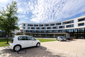 Ein modernes Gebäude mit flachem Dach, zahlreichen Fenstern und einem gepflasterten Eingangsbereich mit geparkten Autos.